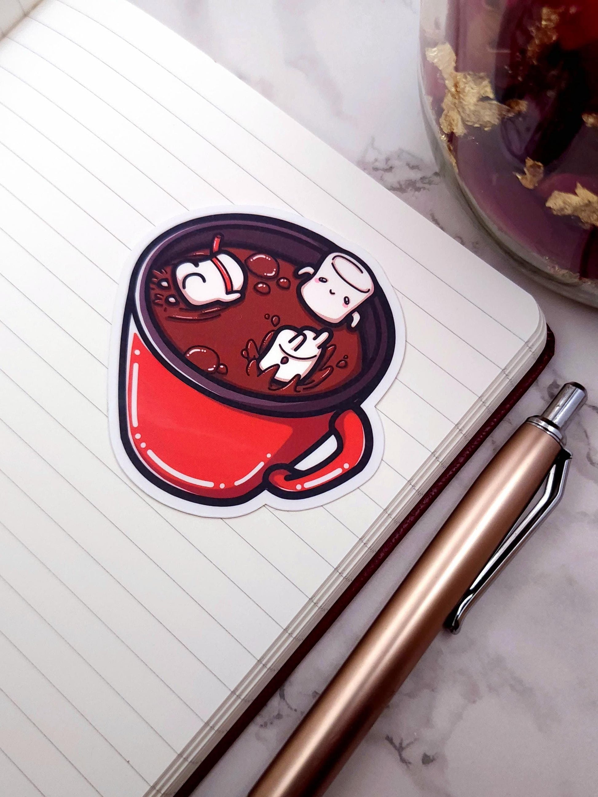 Sticker of a hot chocolate mug with marshmallows on a notebook with a pen next to it.