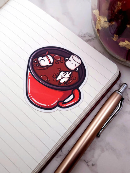 Sticker of a hot chocolate mug with marshmallows on a notebook with a pen next to it.